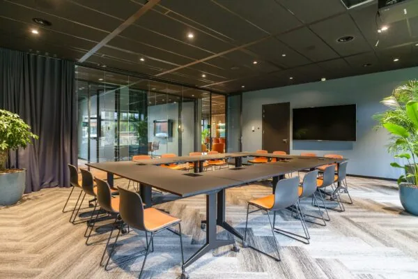 Moderne vergaderruimte aan de George Hintzenweg met U-vormige tafel, oranje stoelen en een flatscreen aan de muur.